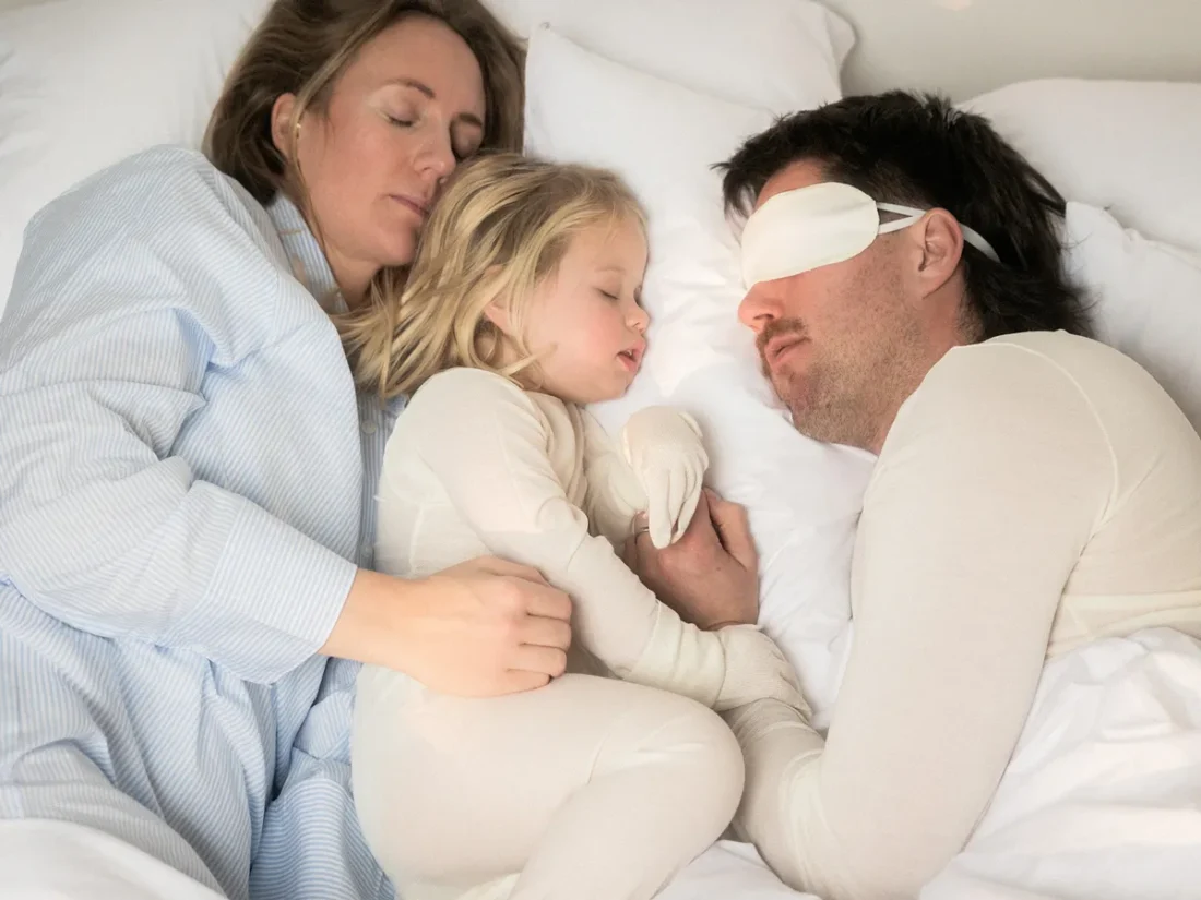 En familj om tre sovandes i en säng. Mamma i blåvitrandig pyjamas håller om dottern som är klädd i helkropps klädsel i DermaSilk. Pappan bär DermaSilk ögonmask och undertröja herr.