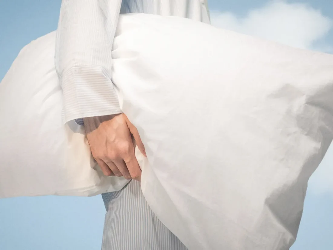 Närbild på en vitt puffig kudde som hålls under armen på en person i blåvitrandig pyjamas. Bakgrunden är klarblå himmel med lätta moln.