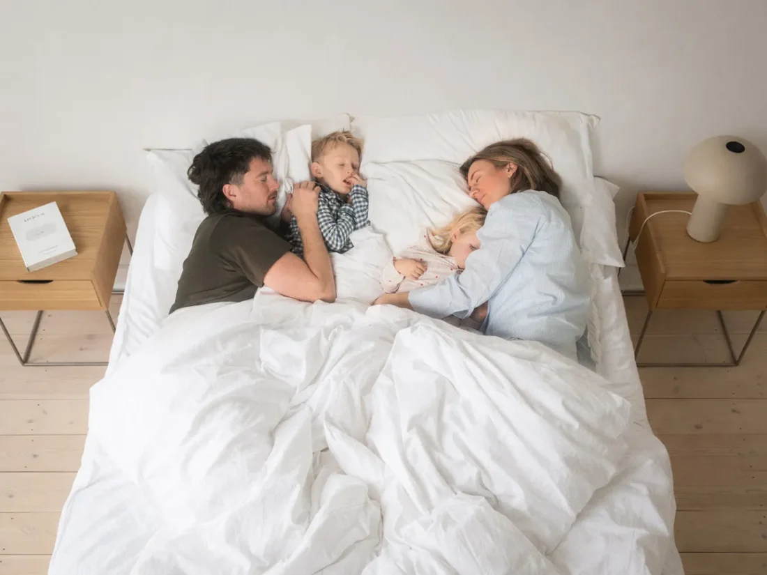 En familj på fyra personer ligger sovande i en dubbelsäng. Pappa och son har mörka pyjamasar, medan mamma och dotter har ljust randiga. Bredvid sängen står två nattduksbord i ek.
